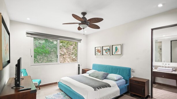 Ein Schlafzimmer mit Bett, Deckenventilator, Fenster mit Blick auf Grünflächen und einem Badezimmer mit Waschbecken und Spiegel.