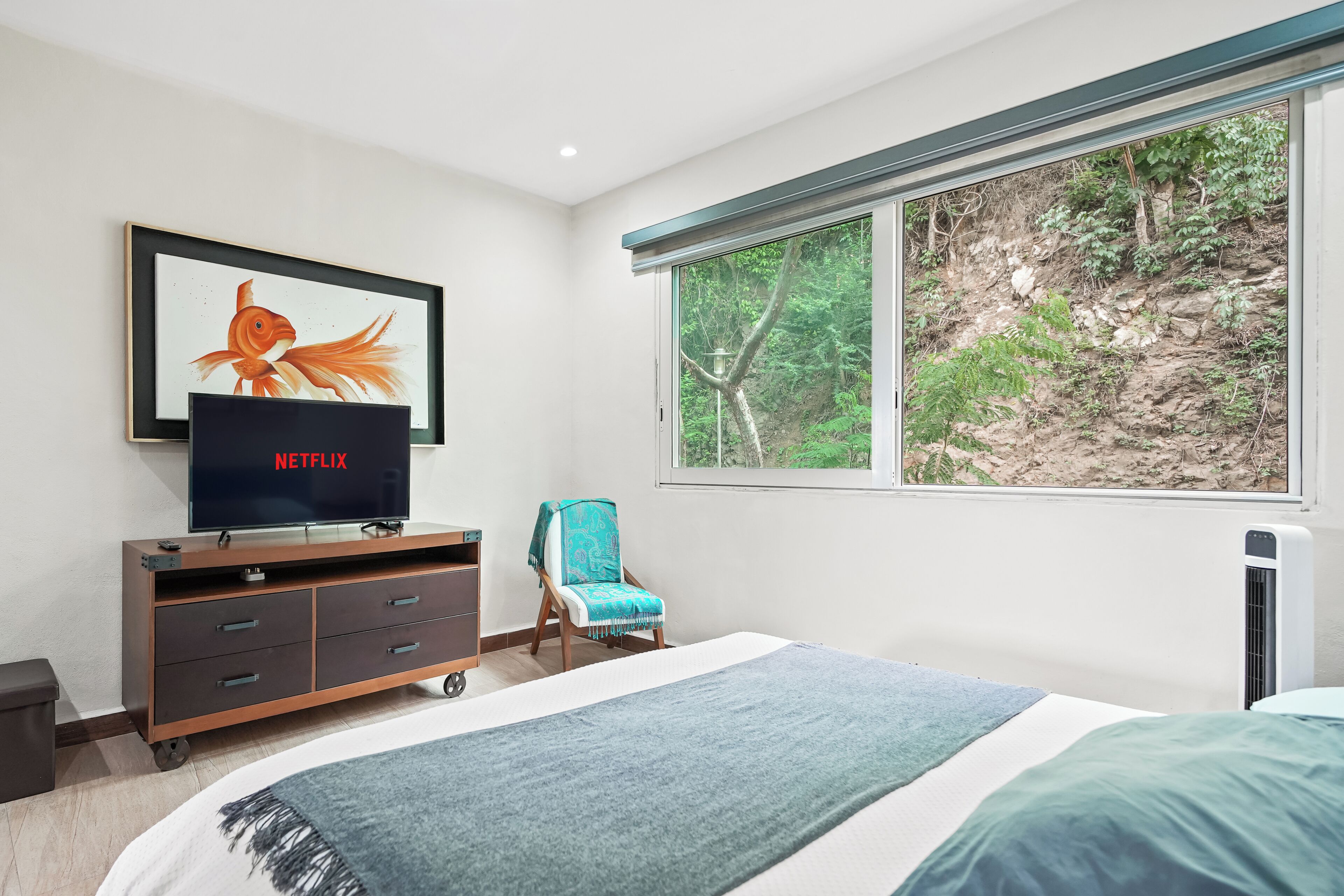 Ein Schlafzimmer mit einem Bett, einem Fernseher auf einer Holzkabine, einem Sessel und einem Fenster mit Blick auf einen felsigen Hügel.