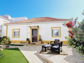 Um edifício branco com telhado ladrilhado, porta frontal e pequeno alpendre com mesa e cadeiras.