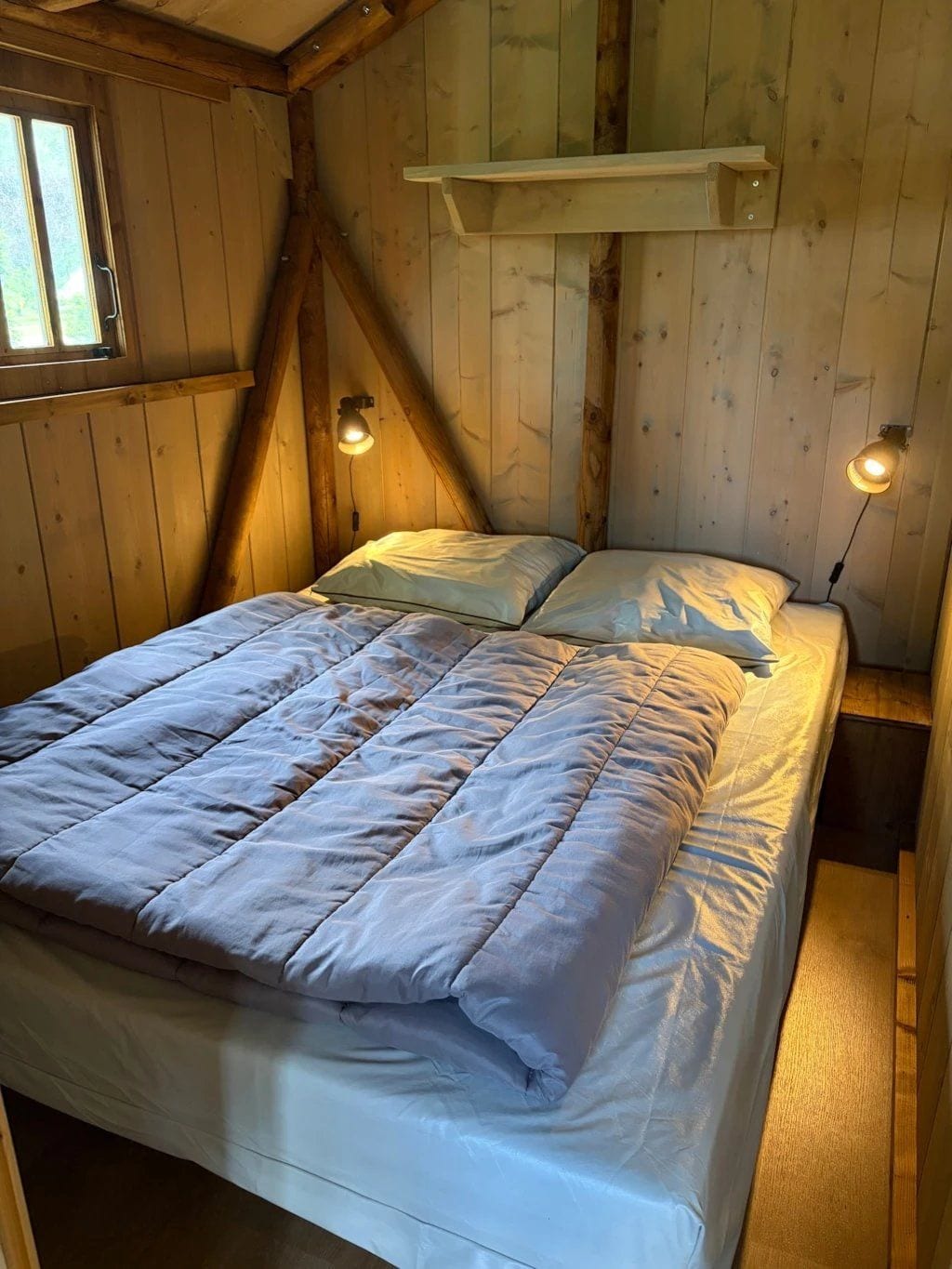 A wooden room with a bed, two wall-mounted lamps, and a window.