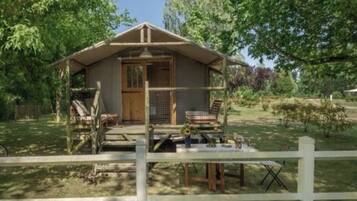 Een klein houten vakantiehuisje met een veranda, omringd door een witte hek en bomen.