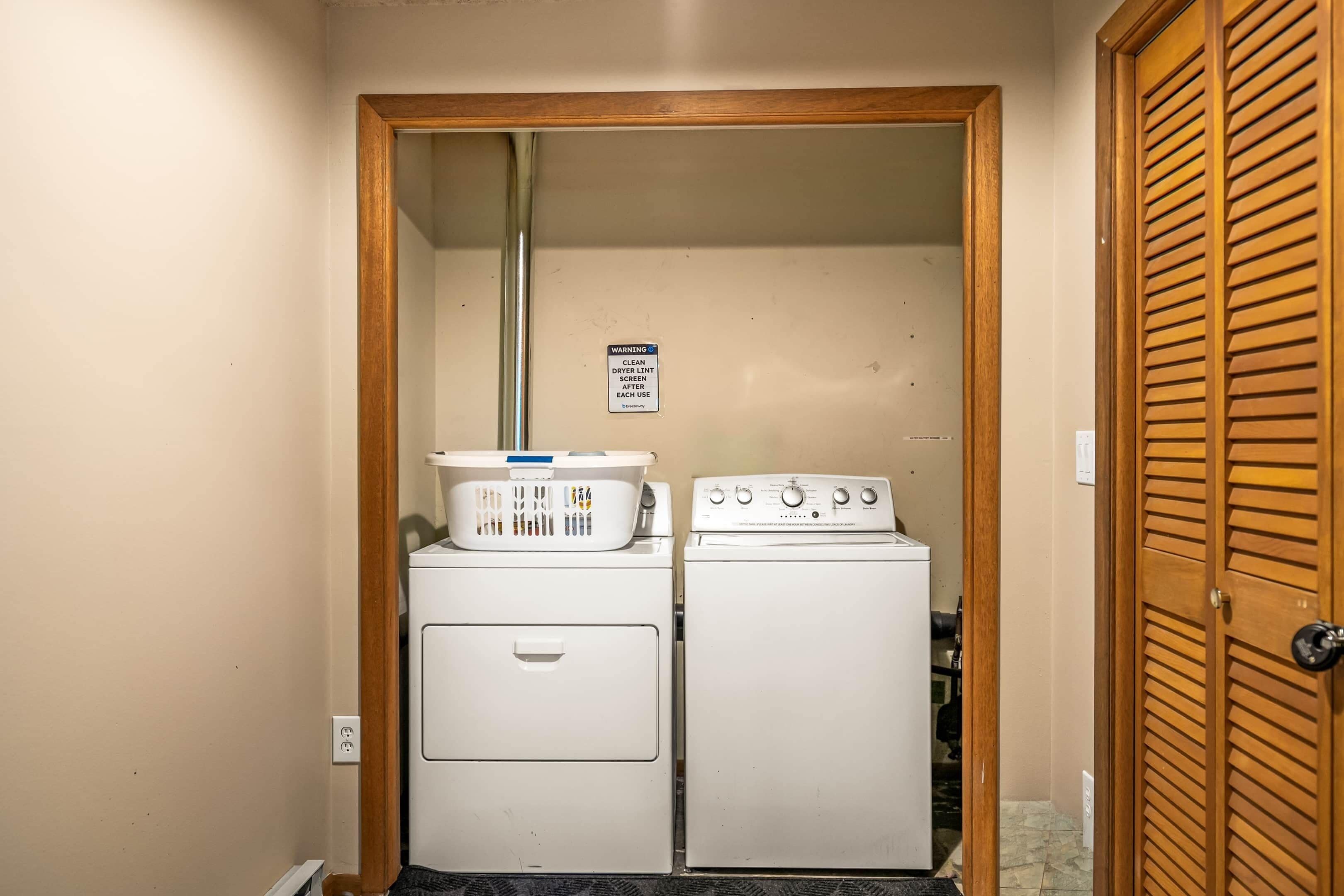 Une buanderie équipée d’un lave-linge et d’un sèche-linge, un panneau d’avertissement sur le sèche-linge et une porte en bois.