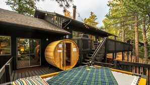 Uma estrutura de sauna de madeira com porta de vidro, um deck de madeira com tapete xadrez e uma escada que leva a um segundo nível.