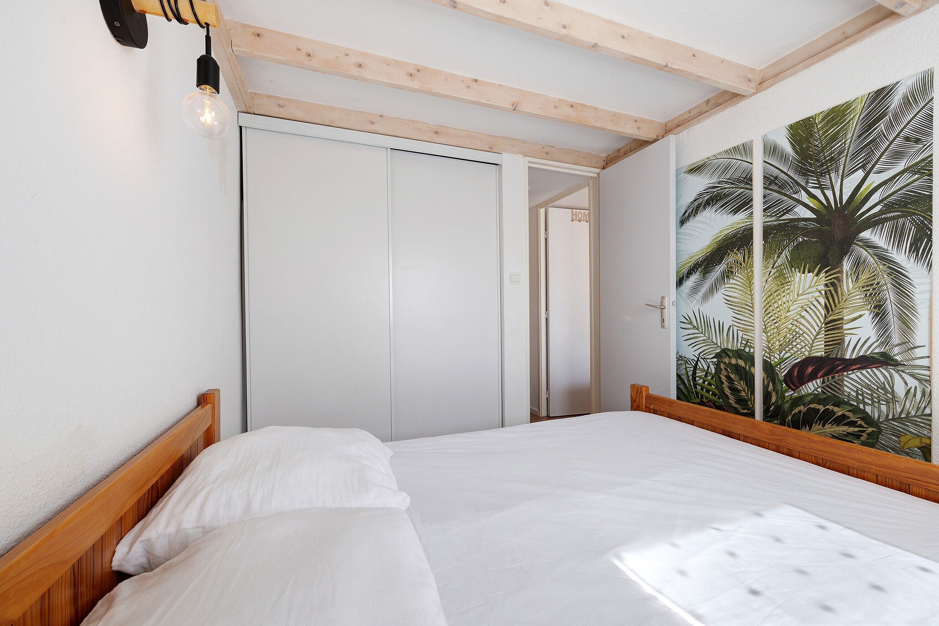 Ein Schlafzimmer mit einem hölzernen Bett, weißer Bettwäsche, einem weißen Kleiderschrank und einer Wand mit tropischen Pflanzen.