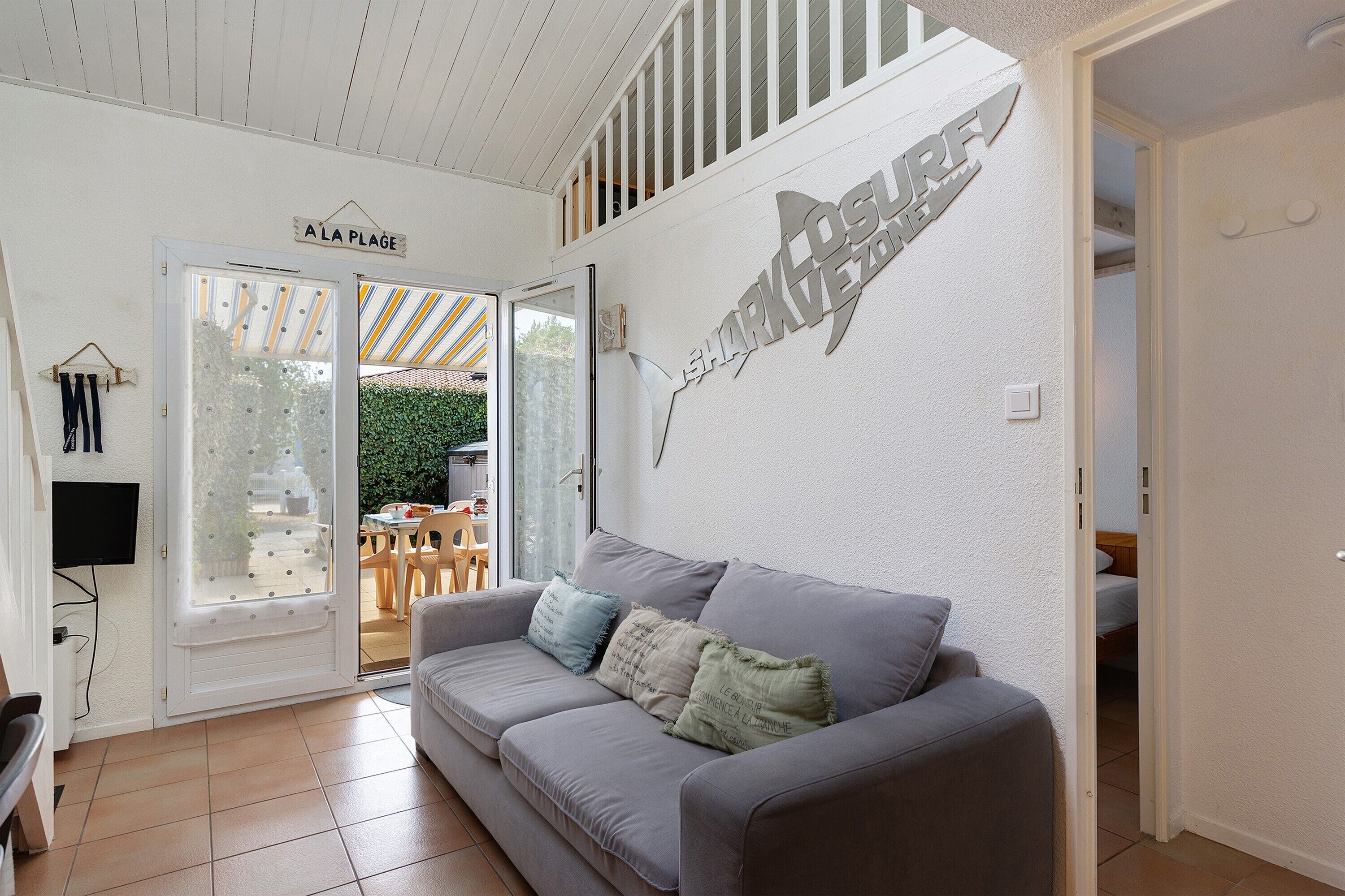 A modern living room with a grey sofa, a flat-screen TV mounted on the wall, and a large window with a view of the outdoors.
