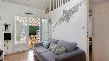 A modern living room with a grey sofa, a flat-screen TV mounted on the wall, and a large window with a view of the outdoors.