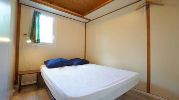 A single bed with a white mattress and blue pillows in a room with wooden beams on the ceiling and a window with a curtain.
