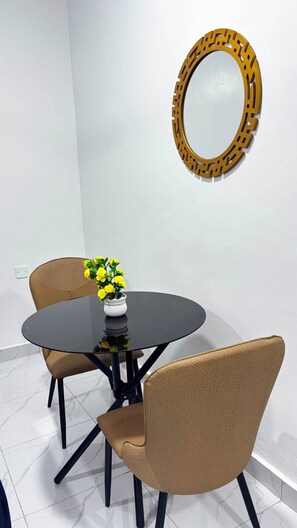Een eetgedeelte met een ronde tafel, twee stoelen, een vaas met gele bloemen en een decoratieve spiegel aan de muur.