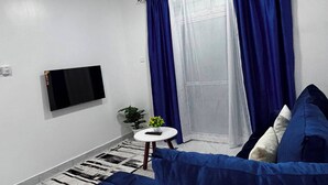 Una habitación con ventilador de techo, televisor de pantalla plana montado, sofá, una mesita redonda con una planta y una ventana con cortinas azules.