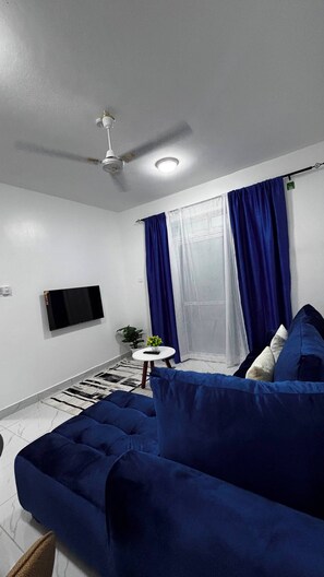 Ein Zimmer mit Deckenventilator, einem fest installierten Flachbildfernseher, einer Couch, einem kleinen runden Tisch mit Pflanze und einem Fenster mit blauen Vorhängen.