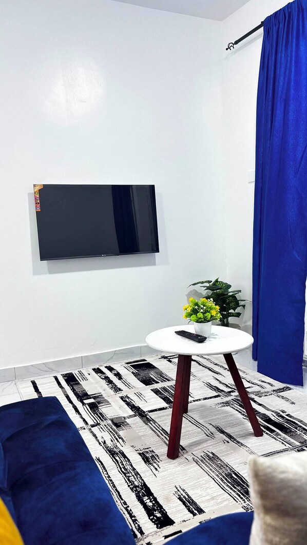 Een moderne woonkamer met een flatscreen televisie aan de muur, een ronde witte tafel met een afstandsbediening, een blauwe bank en een kamerkleed met een patroon.