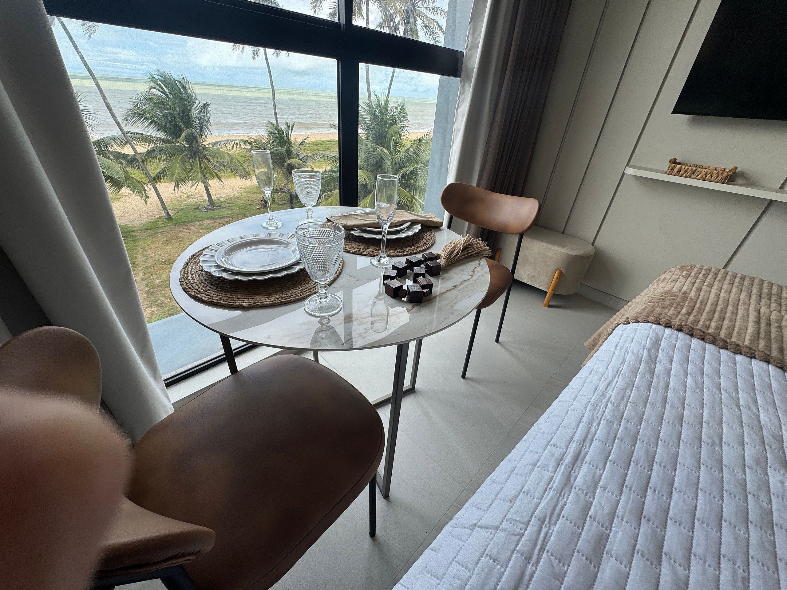 Quarto com cama, mesa de jantar posta com pratos e copos, uma cadeira e vista para a praia e palmeiras através de grandes janelas.