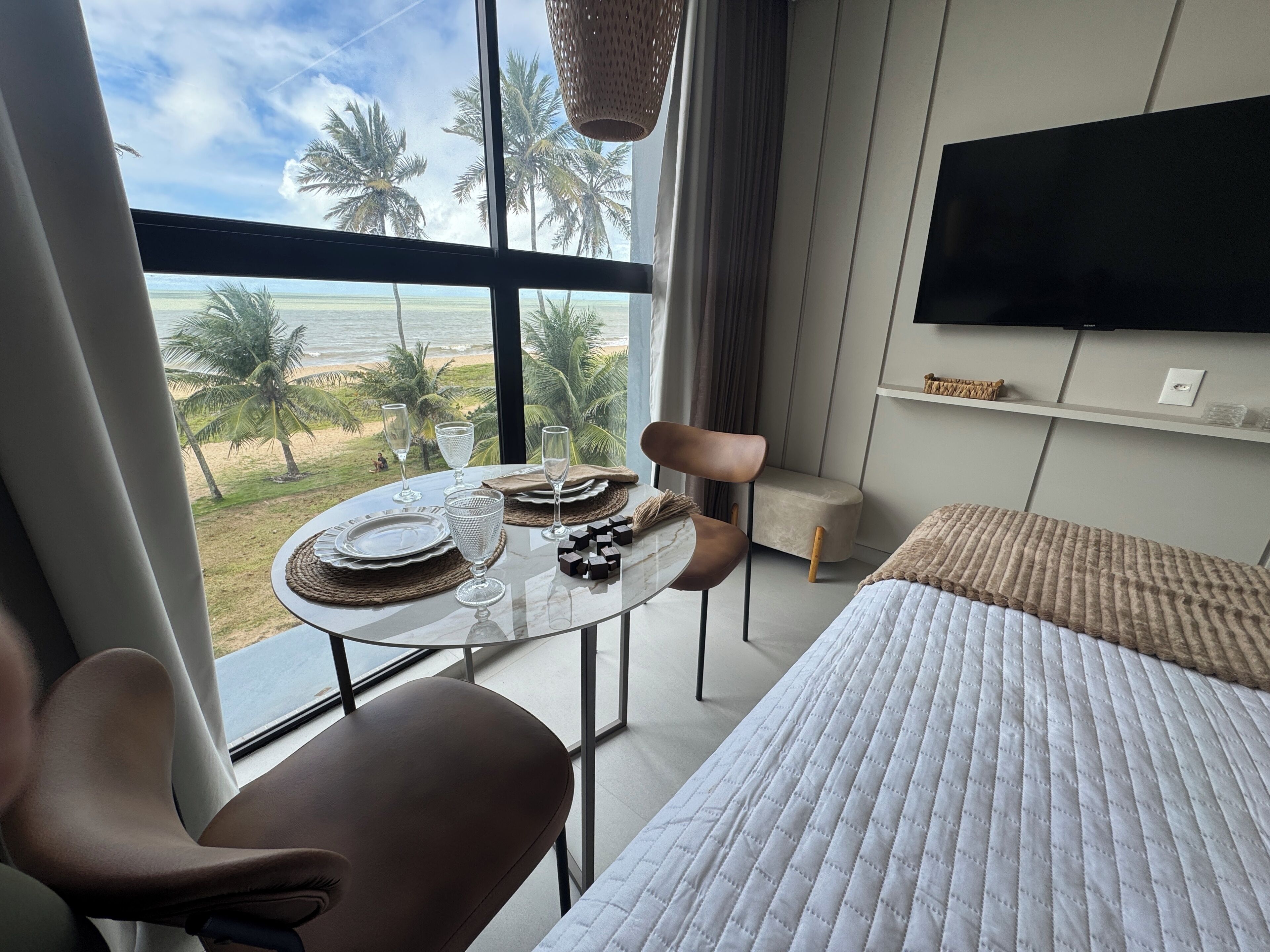 A hotel room with a bed, a dining table with chairs, a TV mounted on the wall, and a view of the beach and palm trees.