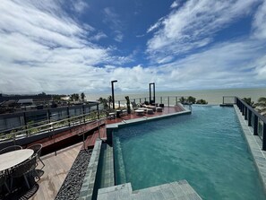 A rooftop infinity pool with a view of the ocean, outdoor seating, and a clear sky. - Breeze Studio Vista Mar Jardim Oceania (João Pessoa)