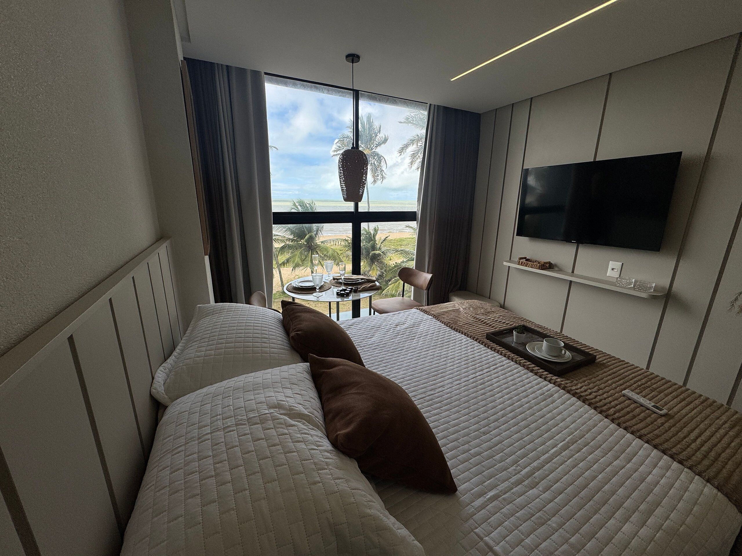 A hotel room with a bed, a TV, a table with a tray, and a view of the beach.