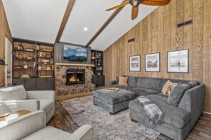 Un salon moderne avec une cheminée en pierre, un grand canapé d’angle gris et un mur en bois orné de tableaux encadrés.