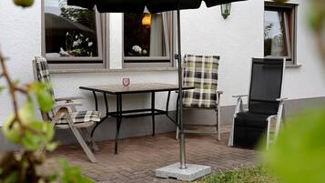 An outdoor patio with a table and chairs, a black umbrella, and a window with a view of the sky.