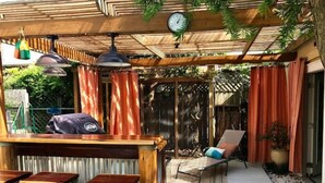 Zona salotto esterna coperta con un bar, sgabelli di legno e una poltrona.