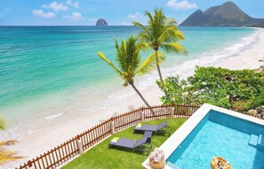 Un área de piscina con sillas de descanso y una valla de madera, con vista a una playa de aguas turquesas cristalinas y una palmera.