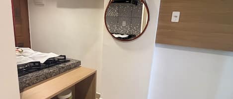 Una habitación compacta con kitchenette, un espejo colgado y una estantería con un recipiente.