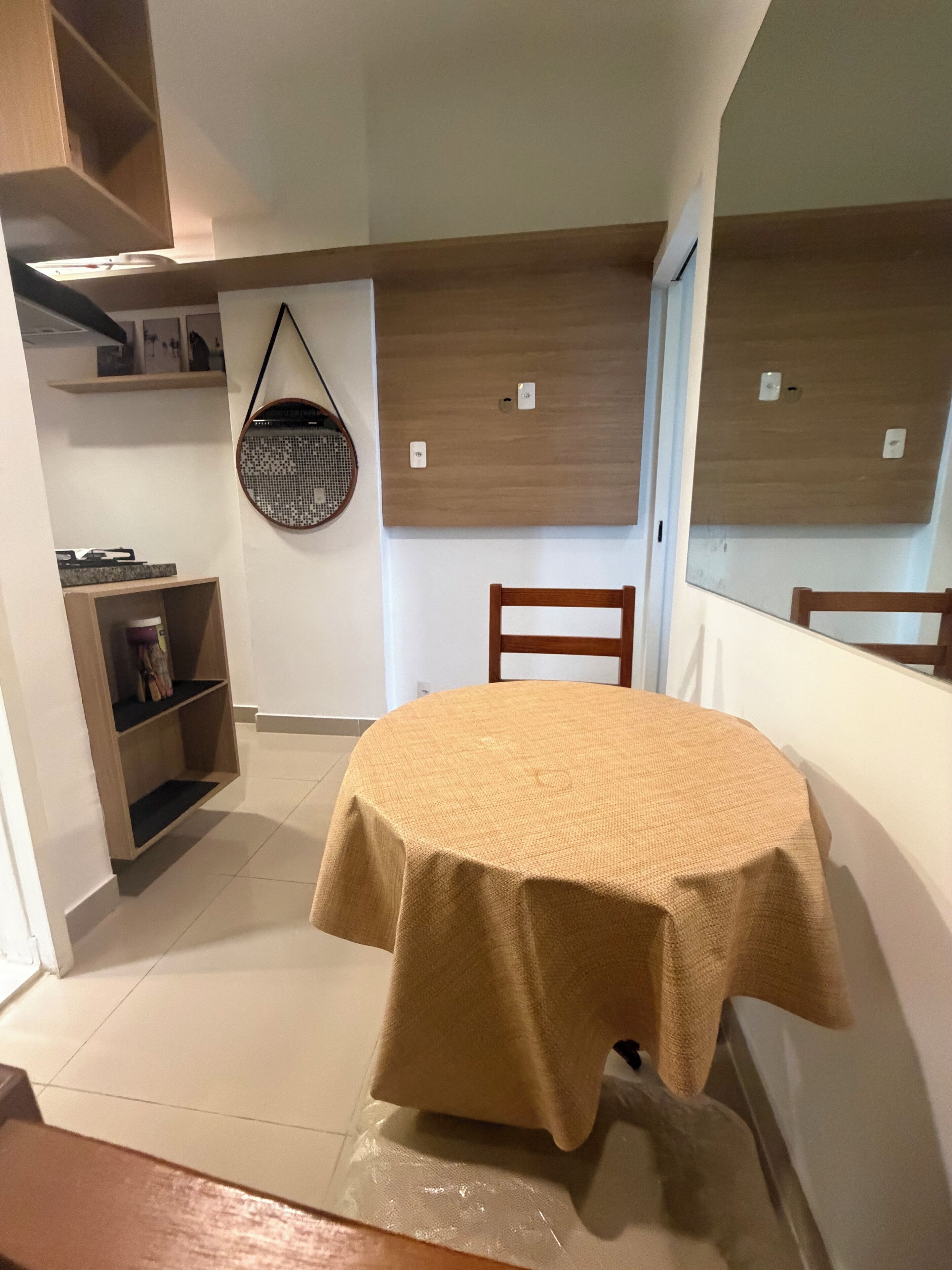 A room with a round table covered by a beige tablecloth, a wooden chair, a wall-mounted basket, and a shelf with items.