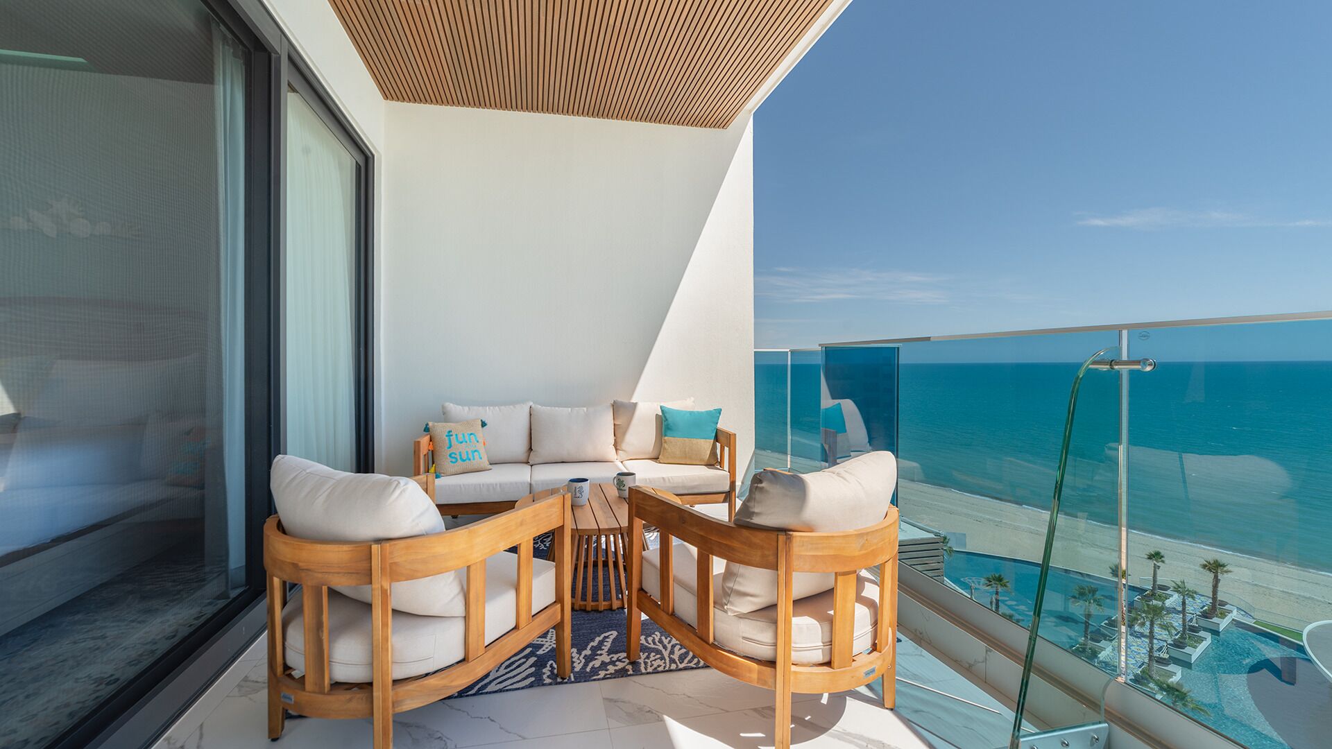 Un balcon avec une table et des chaises en bois, un canapé avec des coussins et une balustrade en verre donnant sur la mer.