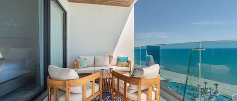 Un balcon avec une table et des chaises en bois, un canapé avec des coussins et une balustrade en verre donnant sur la mer.