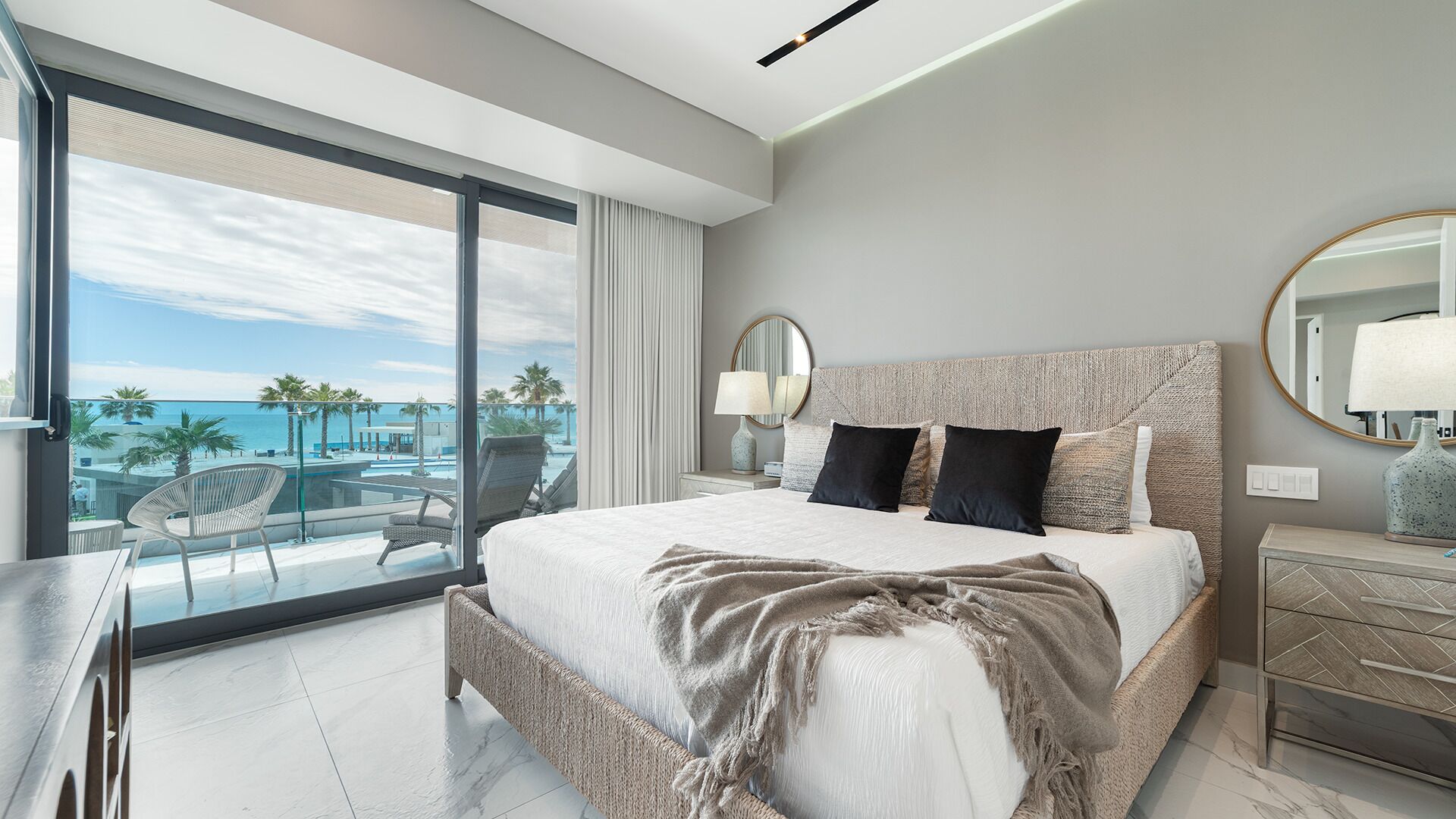 A modern hotel room with a large bed, a balcony with a view of the sea, and a well-lit space.
