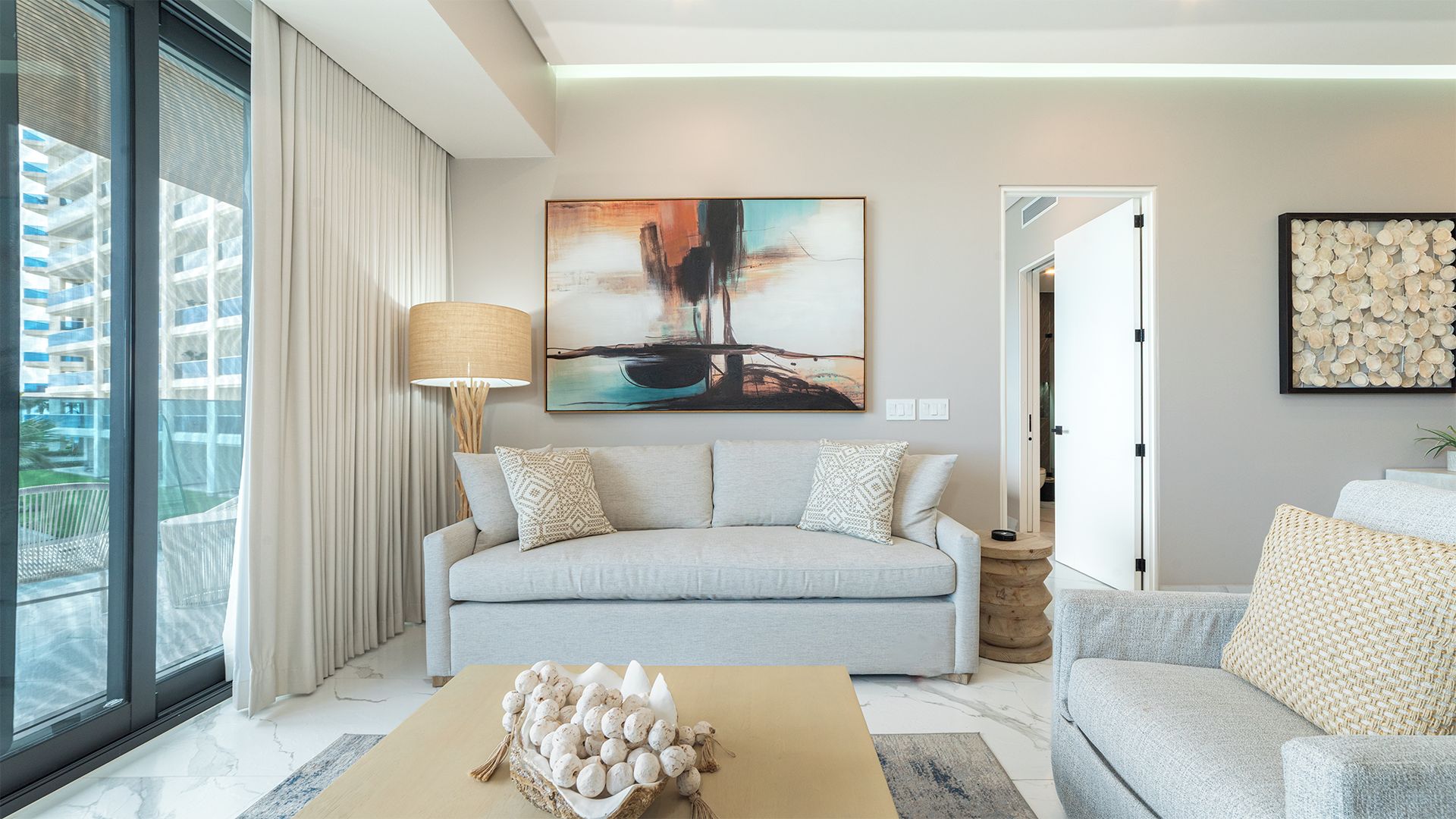 A modern living room with a grey sofa, a coffee table with decorative eggs, a painting on the wall, and a view of a building through large windows.