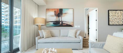 Una sala moderna con un sofá gris, una mesa de centro con huevos decorativos, un cuadro en la pared y vistas a un edificio a través de grandes ventanales.
