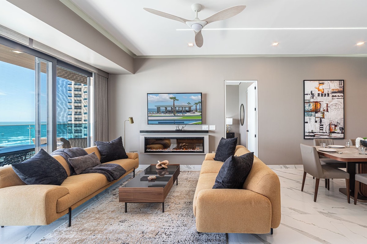 A modern living room with a sofa set, a coffee table, a fireplace, a dining area, and a large window overlooking the ocean.