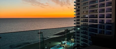 Un balcón con vista al océano al atardecer, un área de piscina y un rascacielos al fondo.