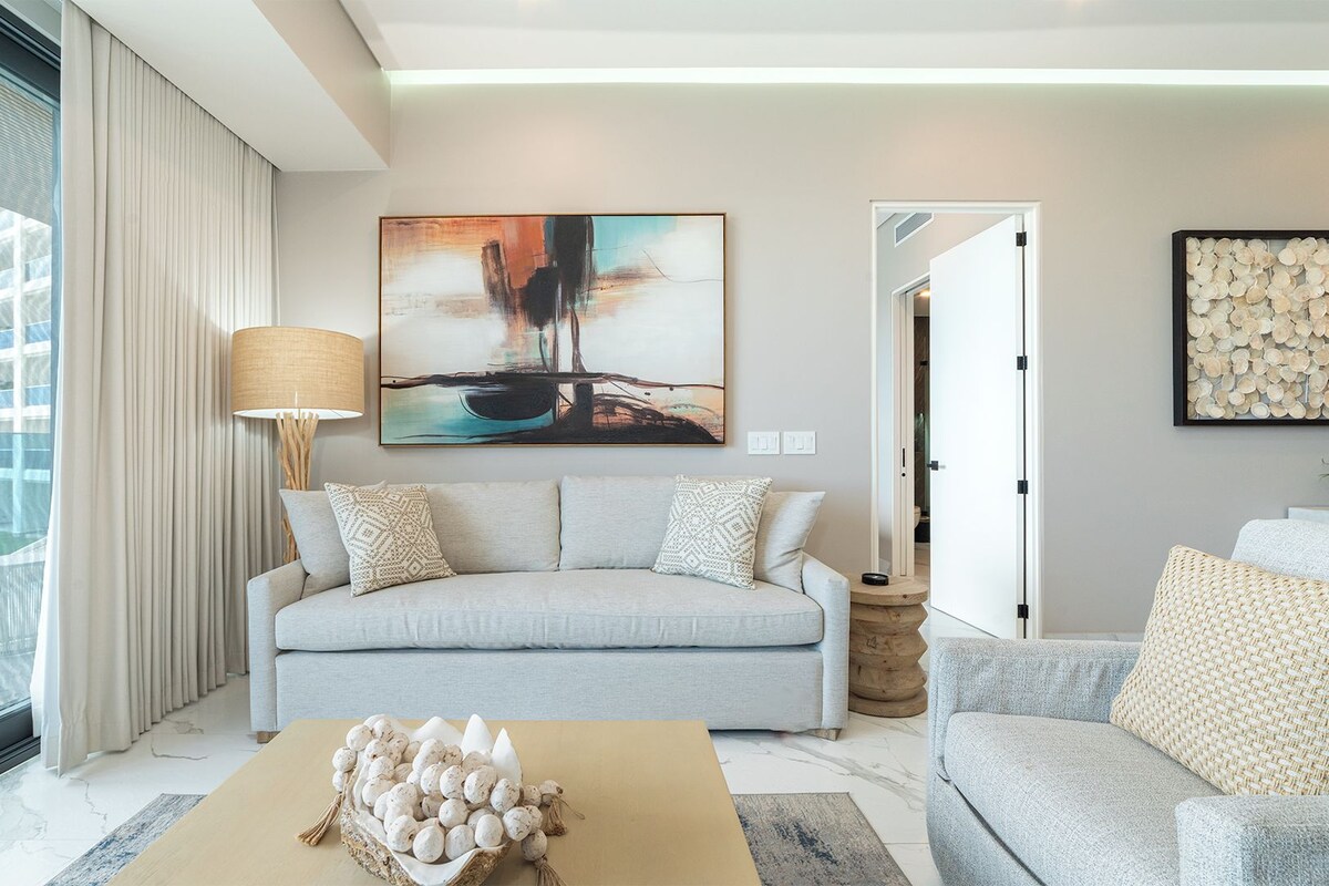 A modern living room with a grey sofa, a coffee table with decorative eggs, a painting on the wall, and a view of a building through large windows.