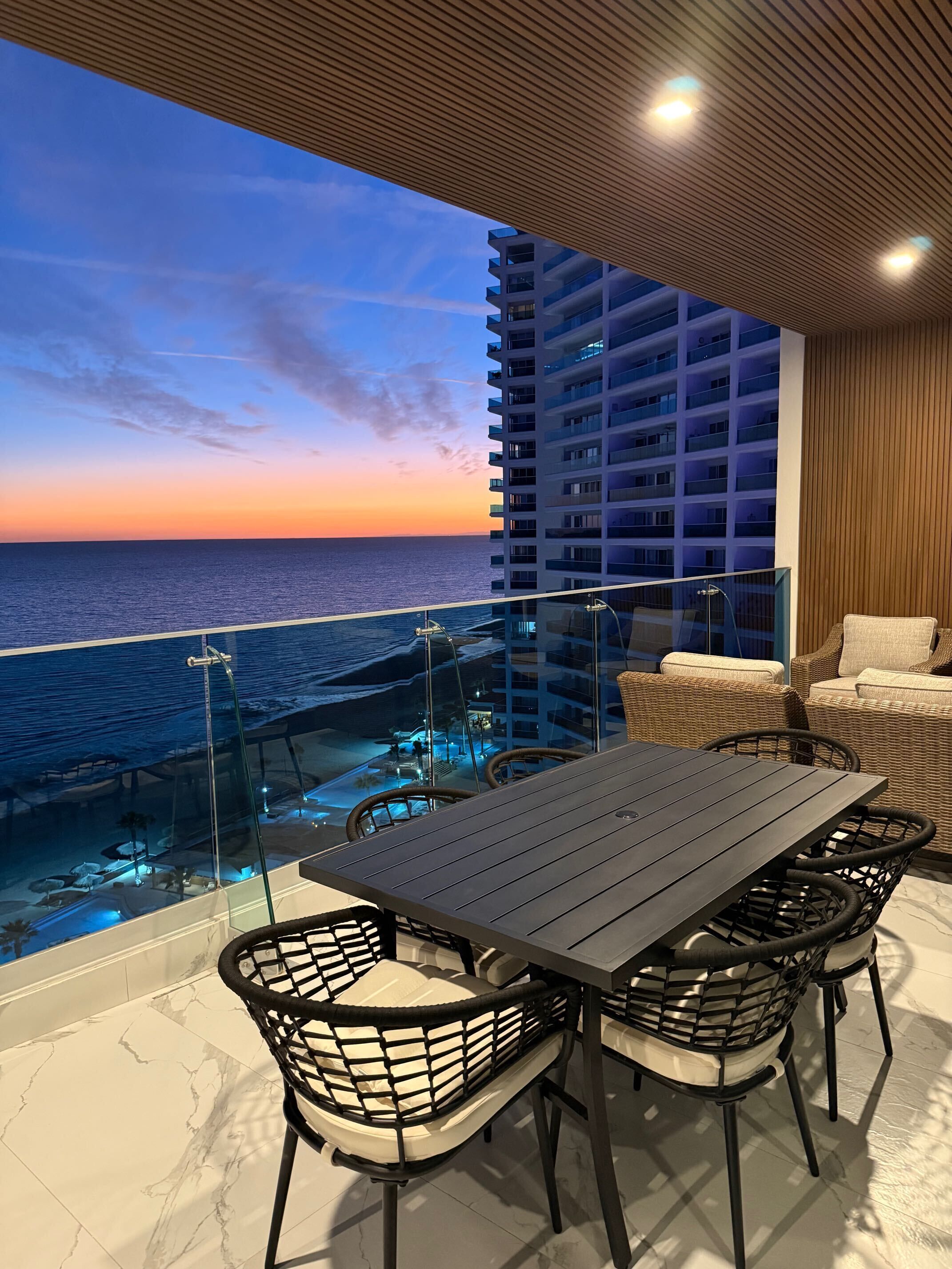 En balkon med udsigt over havet i solnedgang, et spisebord med stole, og en skyskraber i baggrunden.