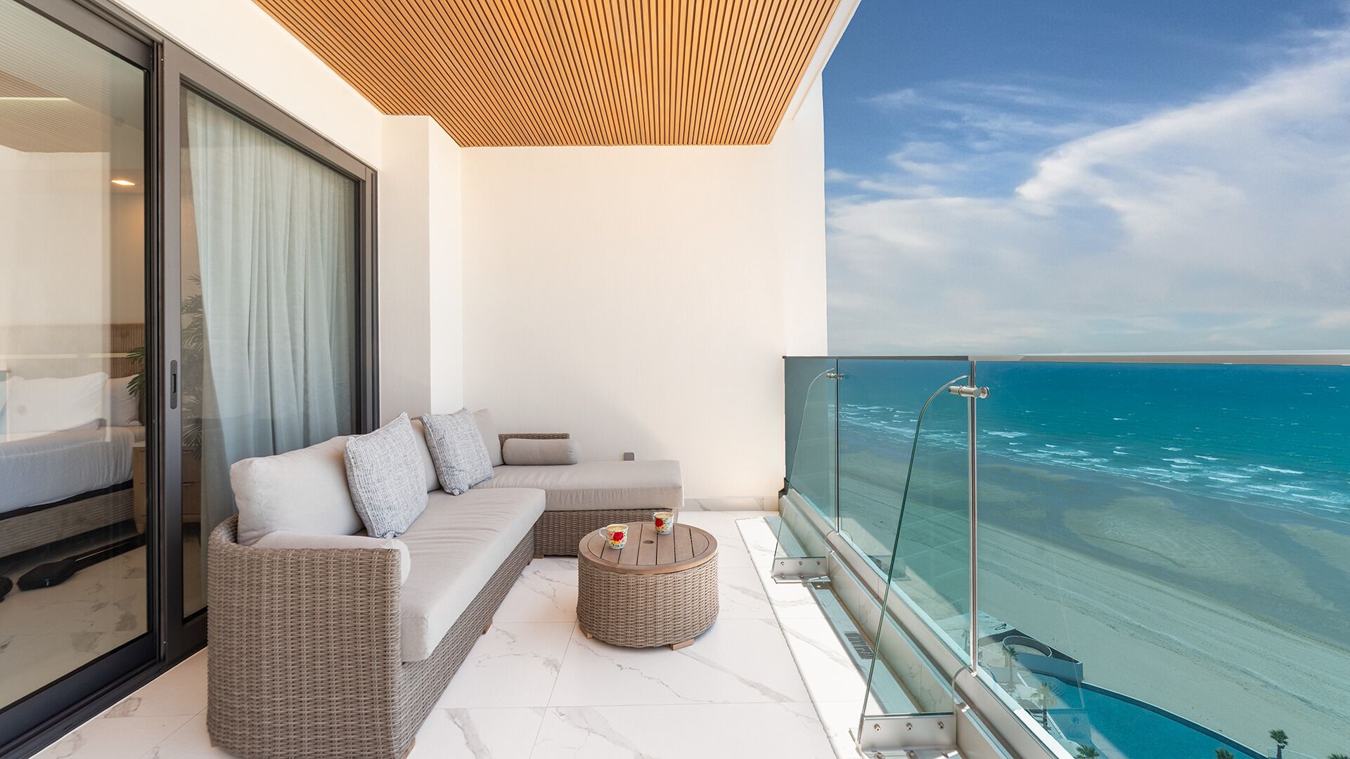 A balcony with a sofa, a round wicker table, and a glass railing overlooking the ocean.