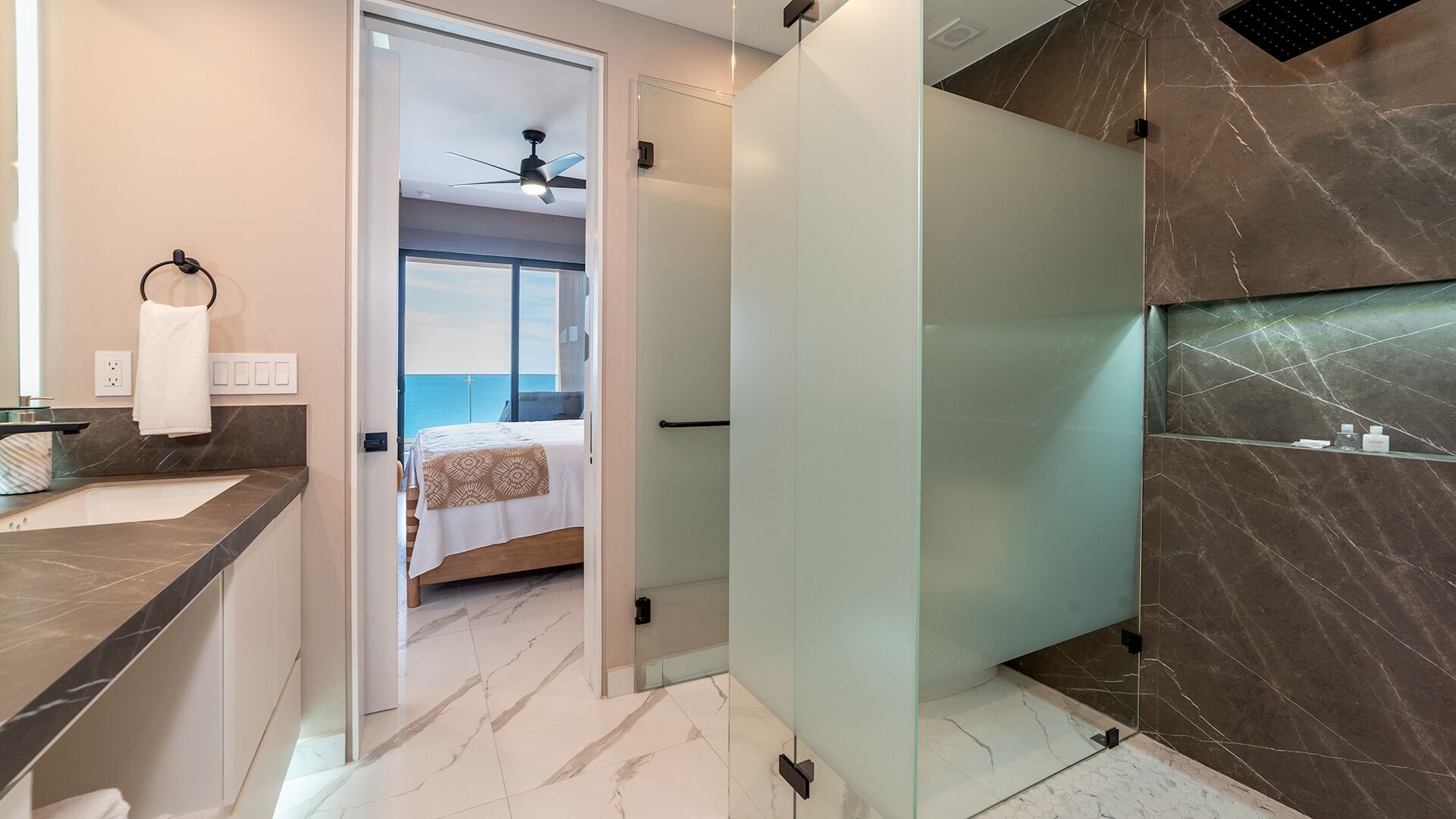 A modern bathroom with a glass partition, a marble floor, and a view of the sea through a door.