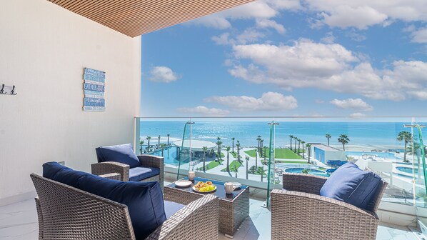 A balcony with wicker furniture, a glass table, and a view of the ocean and resort amenities.