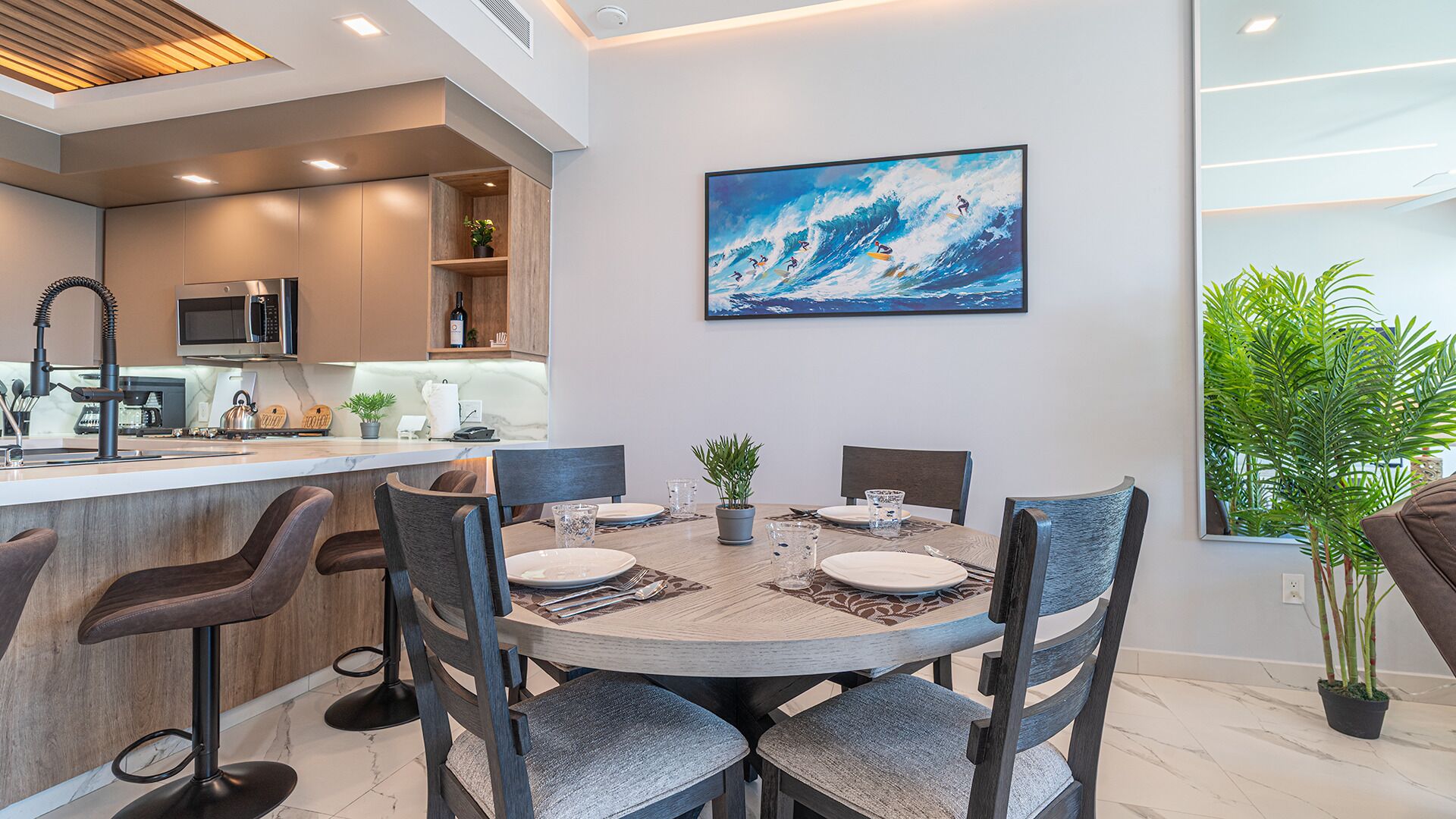 A modern kitchen with a bar area, a dining table set for four, and a large painting of surfers on a wave.