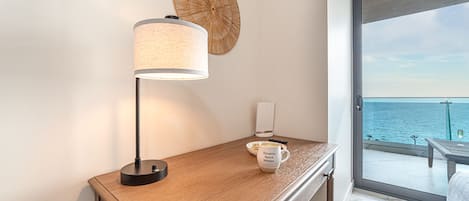 A hotel room with a wooden desk, a lamp, a remote control, a mug, and a view of the sea through a glass door.