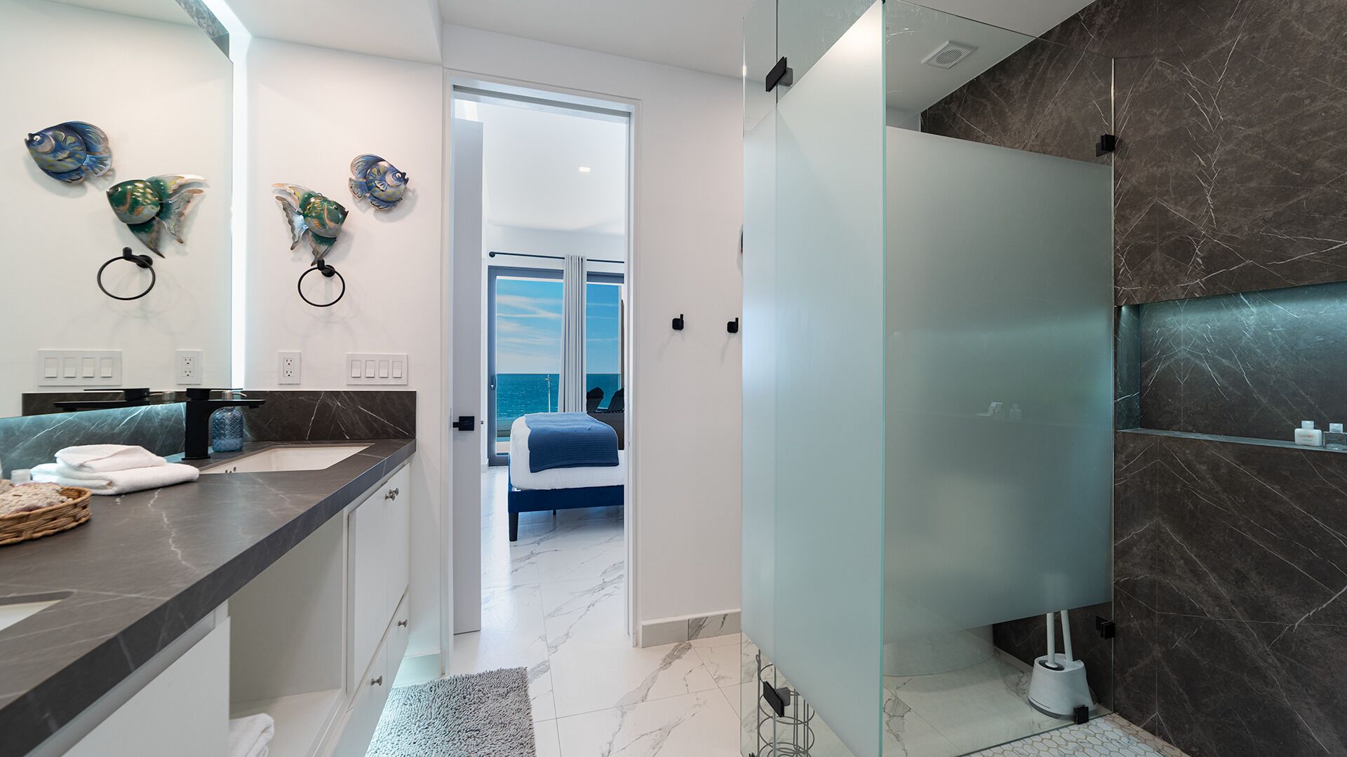 A modern bathroom with a dark marble wall, a glass shower enclosure, and a visible bedroom with a bed and a view of the sea.