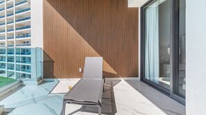 A balcony with a sun lounger, a glass railing, and a view of a modern building with vertical wooden panels. - Encantame Soleil (Puerto Peñasco)
