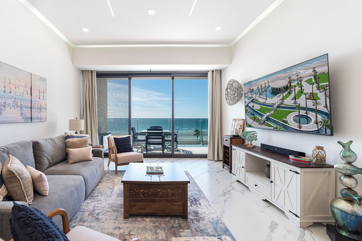 A modern living room with a sofa, coffee table, and a view of the ocean through large windows.