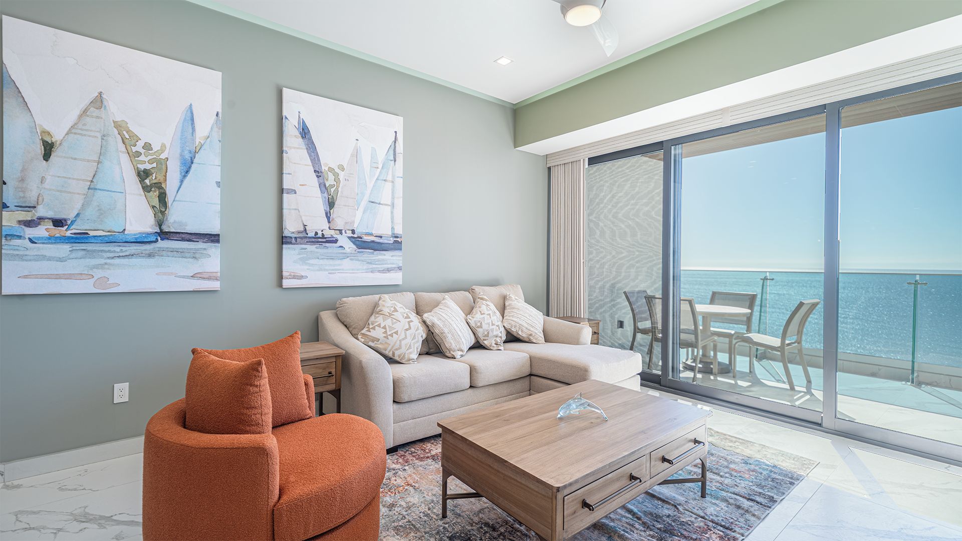 Un salon moderne avec un canapé, une table basse et un fauteuil orange. Deux tableaux ornent le mur, il y a un balcon avec vue sur la mer et une porte vitrée donnant sur l’extérieur.