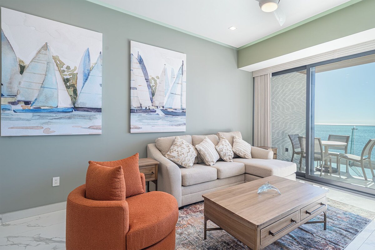 A modern living room with a sofa, coffee table, and orange chair. There are two paintings on the wall, a balcony with a view of the sea, and a glass door leading outside.