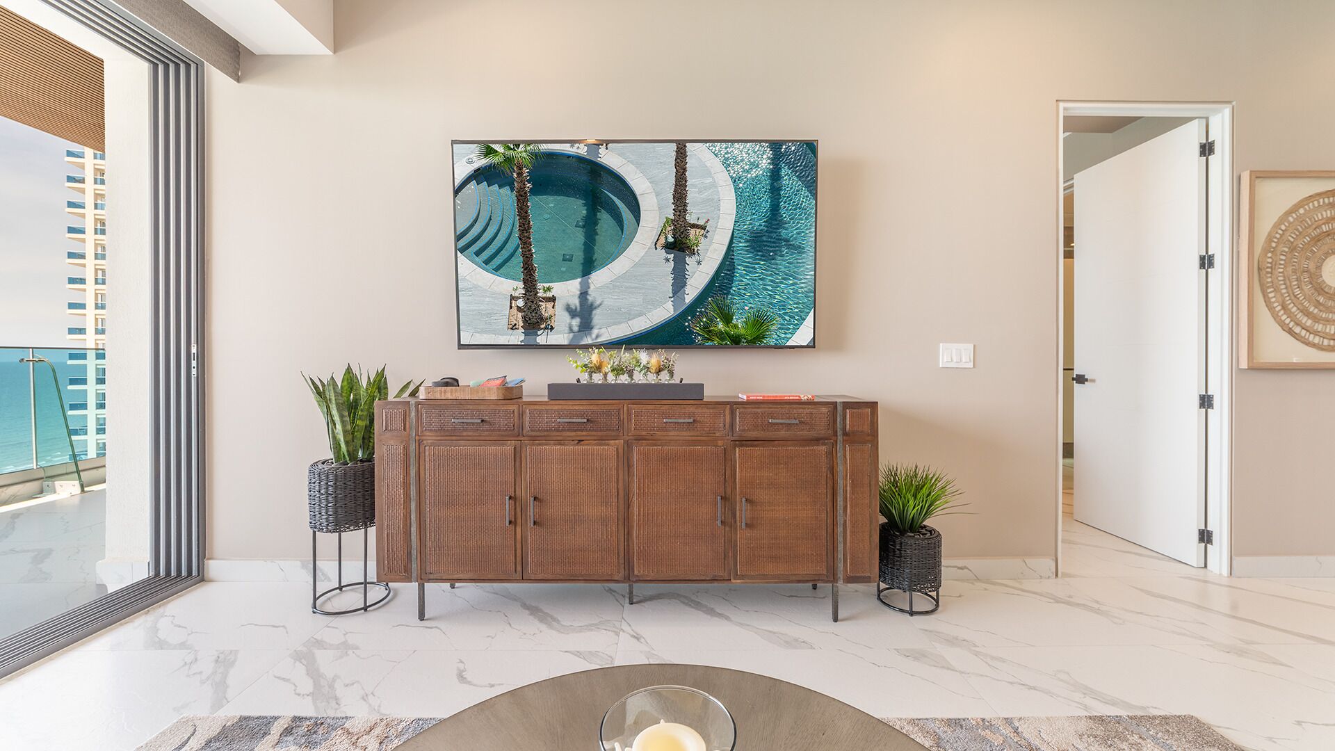 Un interior moderno con una obra de arte grande que muestra una piscina y palmeras, un mueble de madera con cajones, y plantas en macetas.