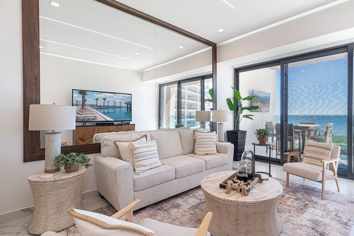 A modern living room with a sofa, coffee table, and chairs, a television, and a large window with a view of the ocean.