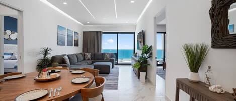 Una sala de estar moderna con un ventanal grande con vista al mar, un comedor con mesa y sillas de madera, y un rincón de descanso acogedor con un sofá.