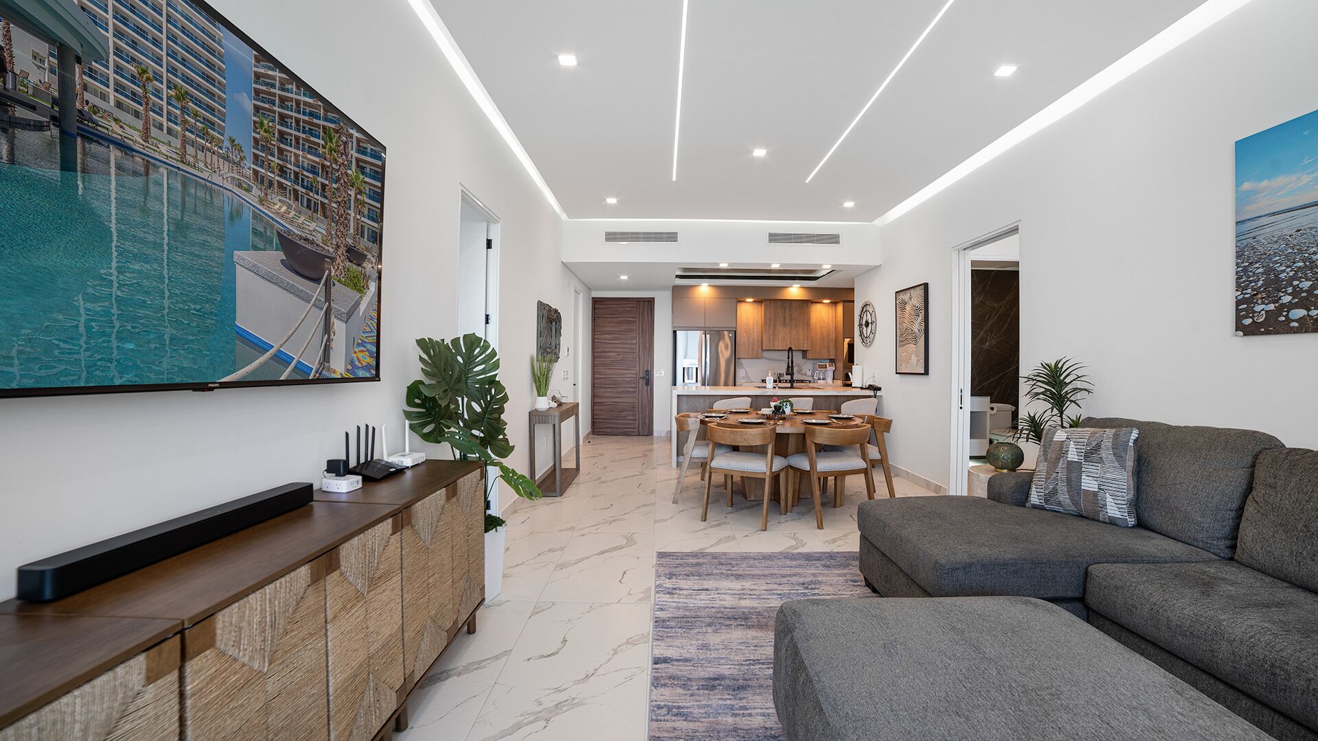 Un salon moderne avec un canapé gris, un coin repas avec une table et des chaises en bois, et une cuisine en arrière-plan.