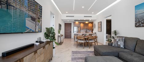 A modern living room with a grey sofa, a dining area with a wooden table and chairs, and a kitchen in the background.