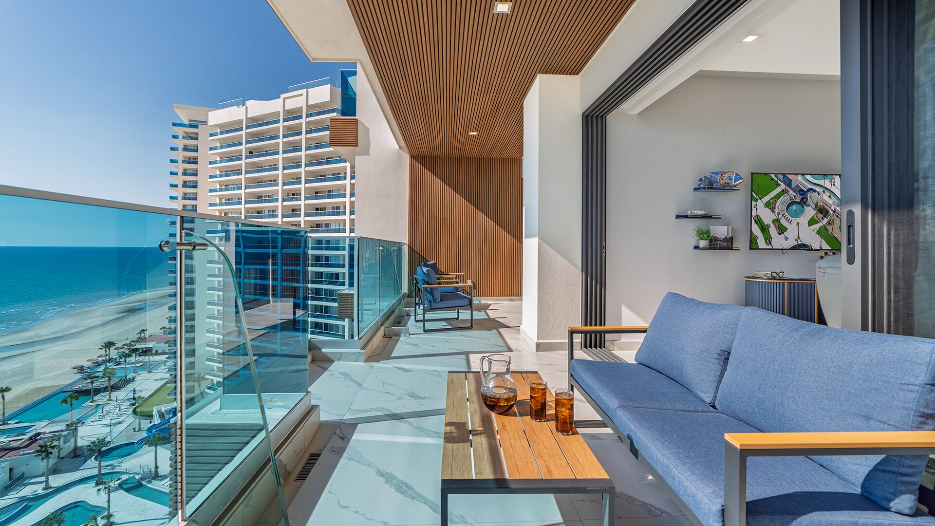 Un balcón con vista a la playa y edificios de hotel, un sofá, una mesa con bebidas y un televisor en la pared.